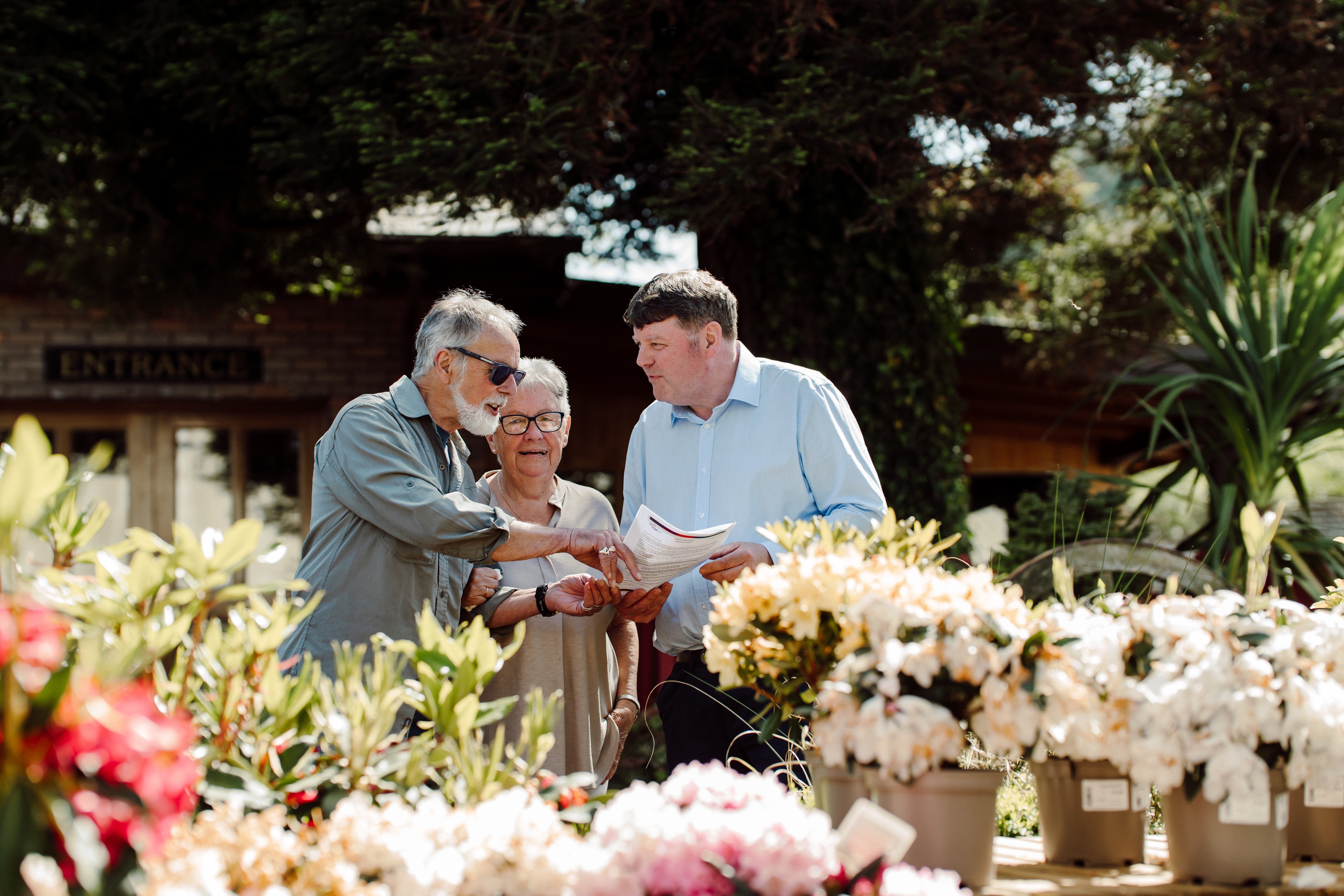 two older people smiling and looking at a sheet of paper with an Age NI staff member with flowers in foreground