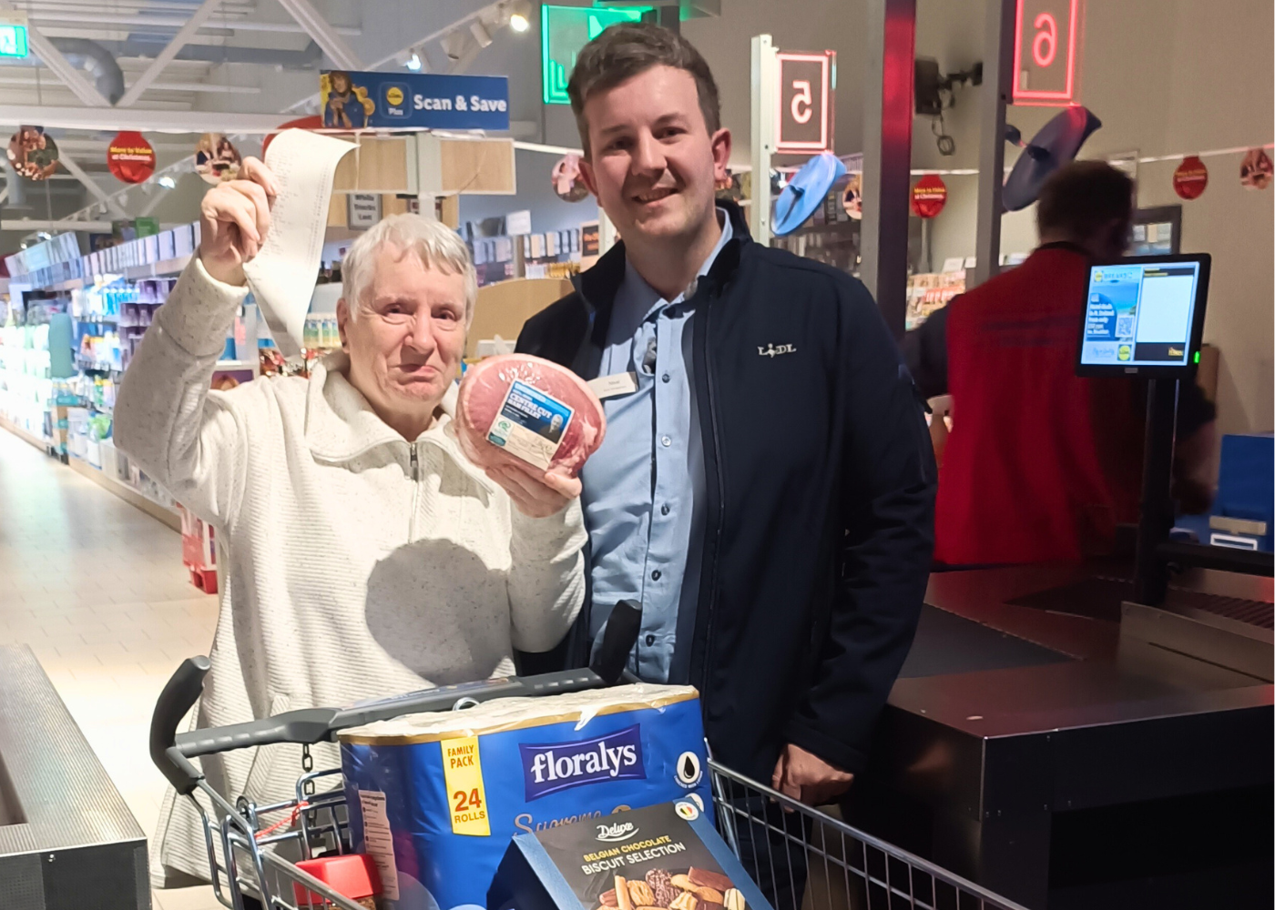 older woman smiling with a full trolley in a Lidl supermarket holding a ham with a member of staff