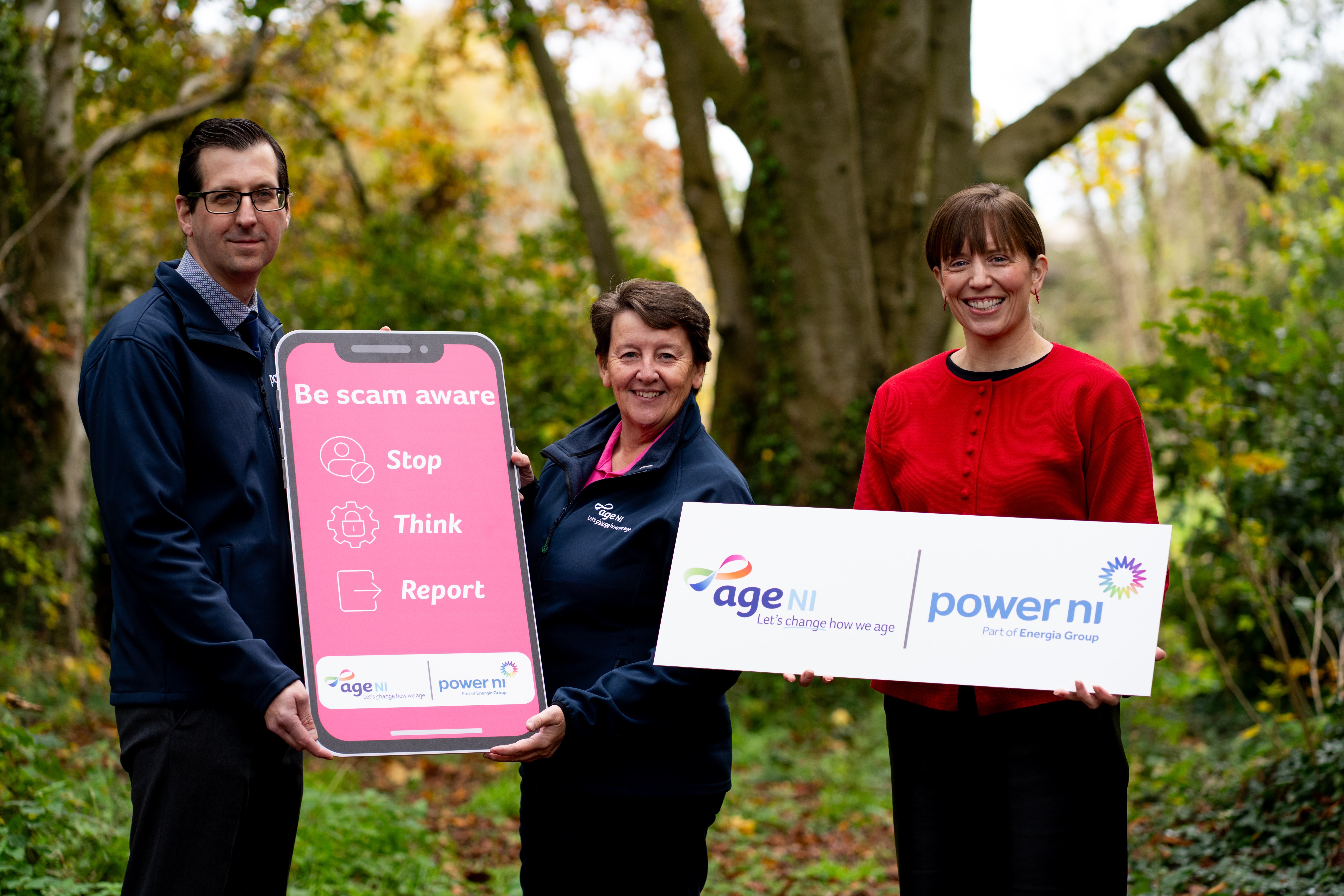 Three people standing outside holding an Age NI/Power NI logo board and a mobile phone shaped prop that reads "Be scam aware: stop, think, report"