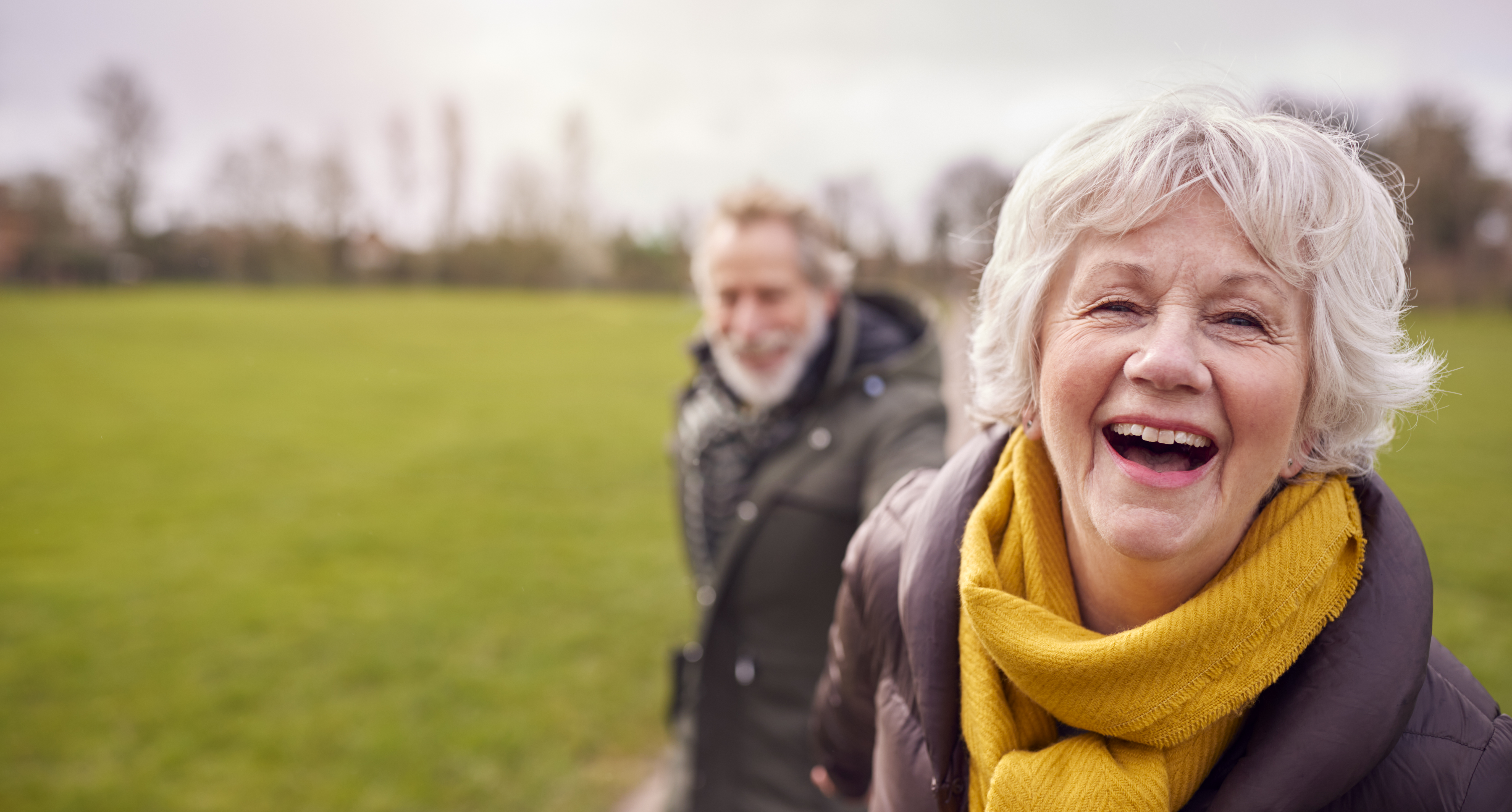 older woman outside wearing a yellow scarf smiling, older man in background