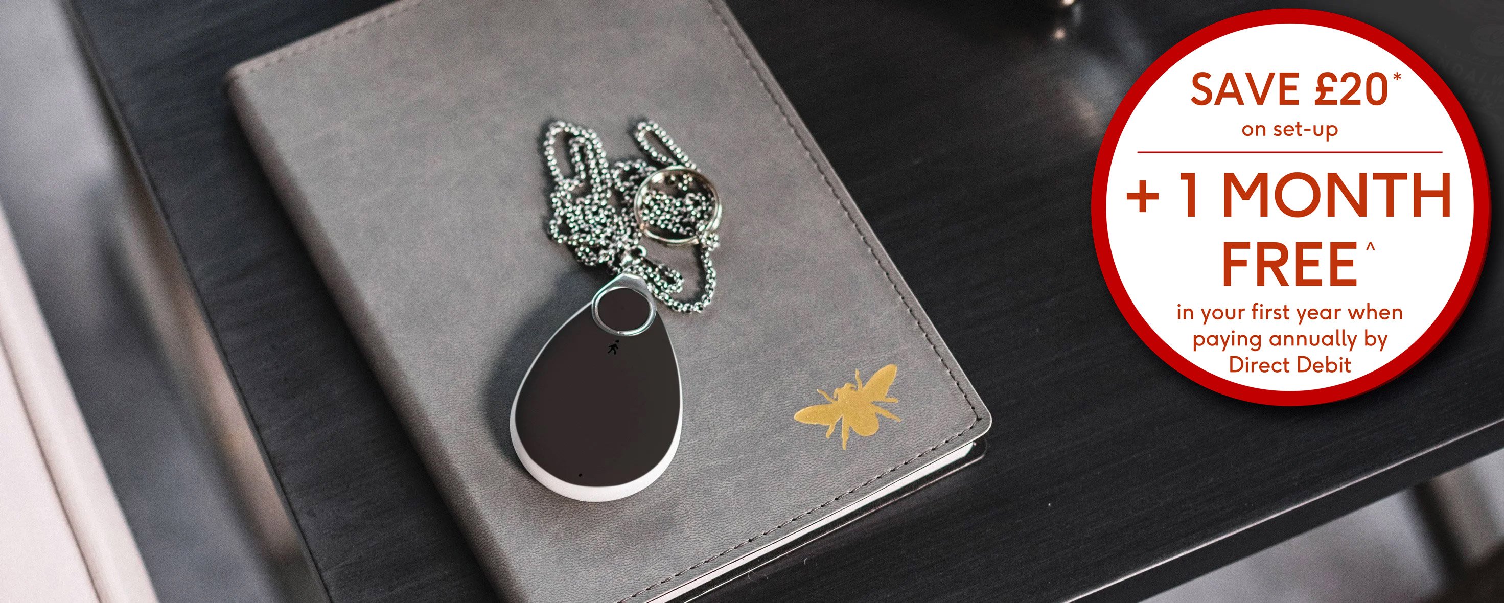 Fall alarm pendant sitting on a notebook on a desk, orange banner in corner "&pound;40 off setup extended for a limited time only"