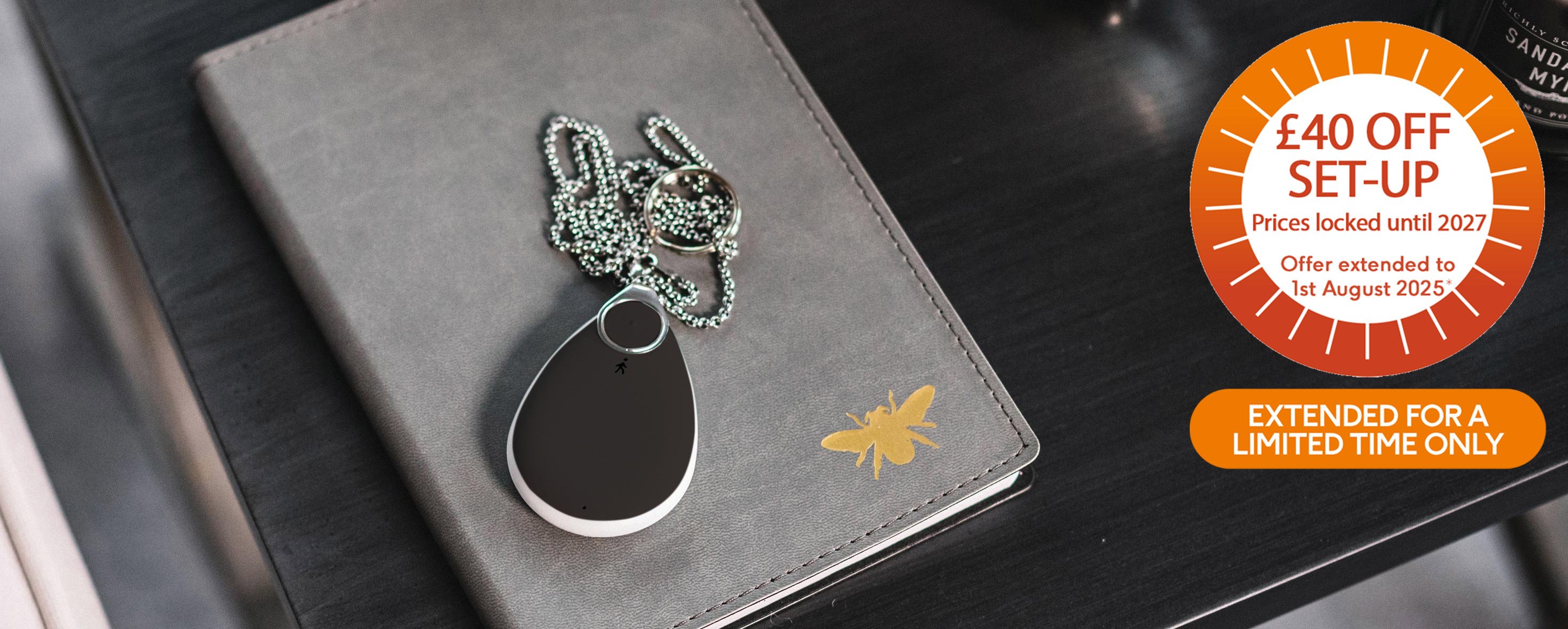 Fall alarm pendant sitting on a notebook on a desk, orange banner in corner "£40 off setup extended for a limited time only"